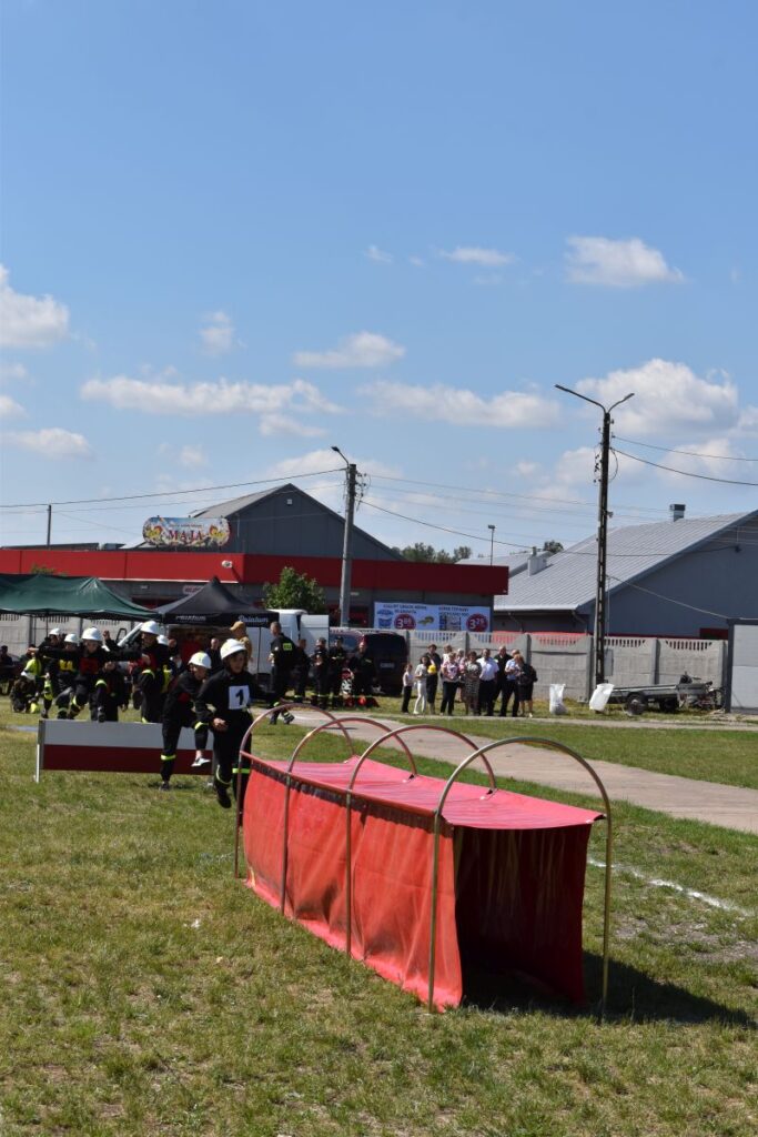 Na obrazku strażacy podzieleni na urządzenia na świeżym powietrzu, używając sprzętu i czerwonych tuneli na trawiastym polu. W tle widoczni są widzowie oraz inne osoby znajdujące się w pobliżu budynków.