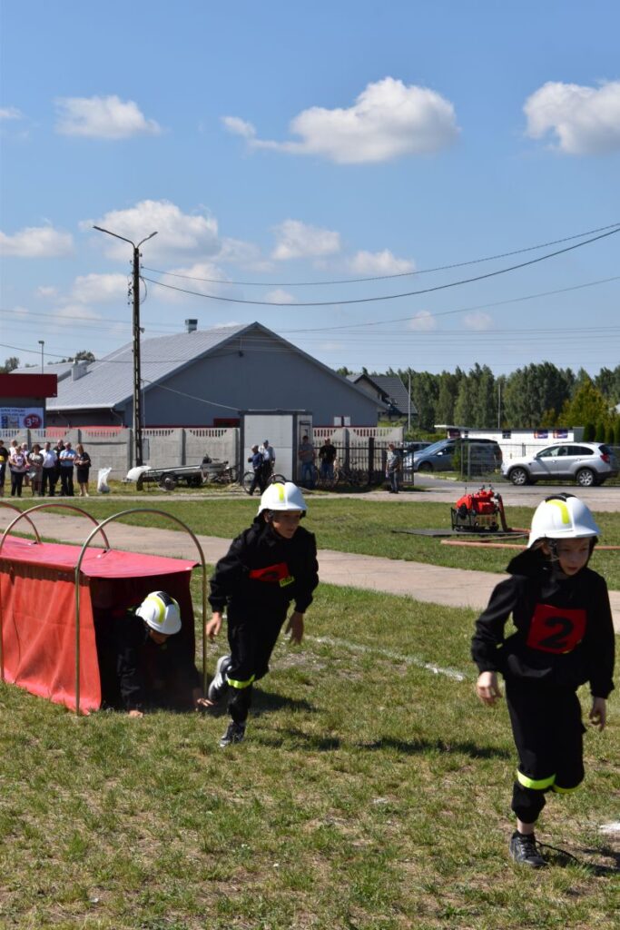 Na obrazku dzieci ubranych w strażackie mundury uczestniczące w terenie torze oddzielne. Jedno dziecko czołgające się przez czerwony tunel, podczas gdy dwaj inni biegają w pobliżu.