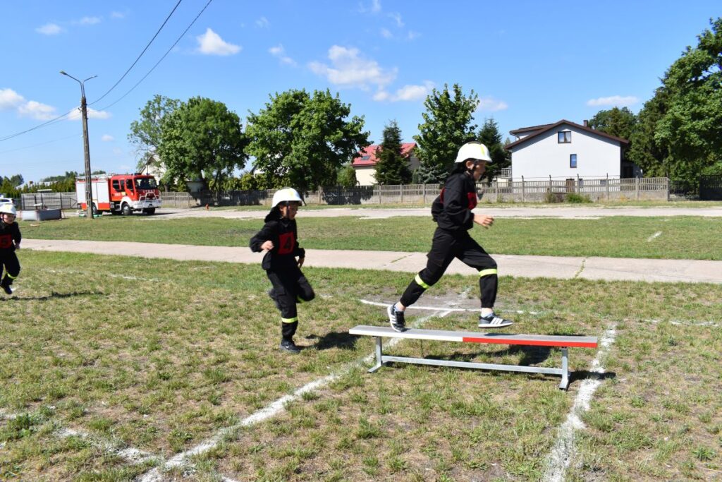 Dwaj strażacy w strojach treningowych biegną po trawiastym polu, z czymkolwiek przeskakuje poziom poprzeczkę. W tle widoczny jest trzeci zbiornik oraz wóz strażacki.