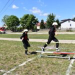 Dwaj strażacy w strojach treningowych biegną po trawiastym polu, z czymkolwiek przeskakuje poziom poprzeczkę. W tle widoczny jest trzeci zbiornik oraz wóz strażacki.