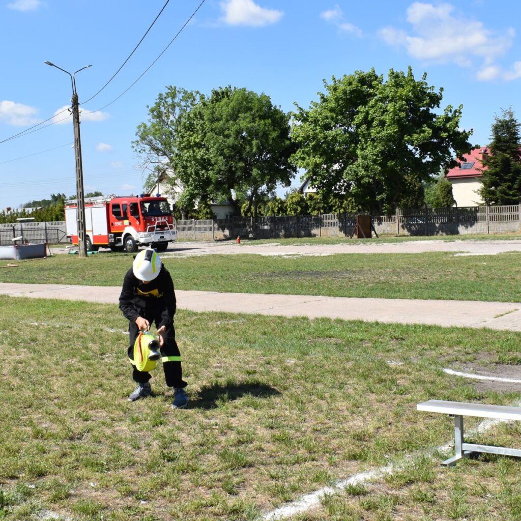 Na obrazie czarnym mundurze i białym hełmie klęczy na trawie obok ławki, a w tle widać wóz strażacki oraz drzewo. Tło własne miejsce otwarte, możliwe, że park lub skwer.