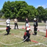 Na obrazku ludzi w mundurach strażackich, wyodrębniających udział w działaniu przeciwpożarowym na świeżym powietrzu, używających węży niszczyzych oraz celów. W tle znajduje się drzewo i dodatkowi uczestnicy szkoleń.