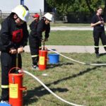 Na obrazku dzieci ubrane w strażackie mundury obsługujące pompy wodne na świeżym powietrzu. W tle widać inne dzieci oraz osobę dorosłą, którzy przypatrują się wydarzeniu.