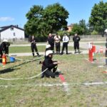 Na obrazku strażacy uczestniczyli w ćwiczeniach na świeżym powietrzu, używając węży do celowania w panelach. Grupa obserwatorów, monitorowane w mundurach, obserwujące działania i notatki.