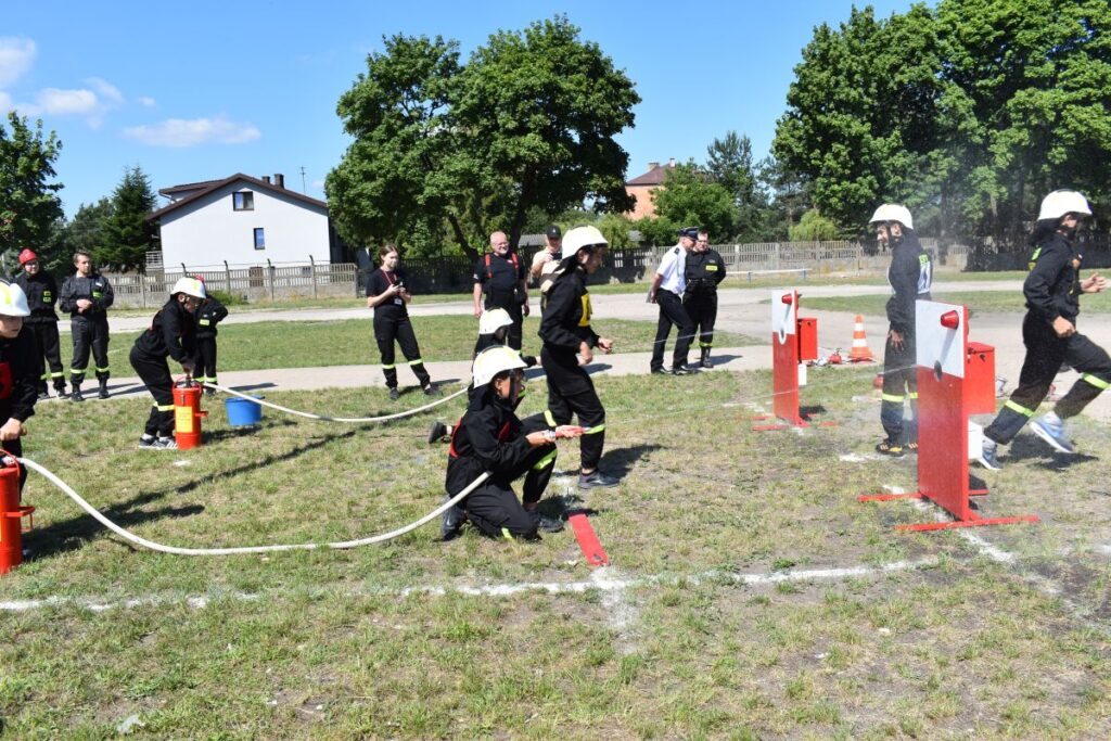 Na obrazku strażacy uczestniczą w ćwiczeniach, obsługując węże i strumienie wody na cele cele. Scena ma miejsce na otwartym terenie z trawą, a w tle są drzewa oraz dostępne.