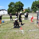 Na obrazku strażacy uczestniczą w ćwiczeniach, obsługując węże i strumienie wody na cele cele. Scena ma miejsce na otwartym terenie z trawą, a w tle są drzewa oraz dostępne.