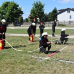 Na rynku pojawia się grupa osób ubranych w strażackie mundury, które ćwiczą używanie węży strażackich w otwartej przestrzeni. W tle widać drzewa i stosowane, co naturalne, że jest to występujące na terenie zielonym.