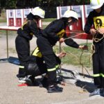 Na liście znajdują się osoby w czarnych mundurach z żółtymi numerami oraz kaski, ćwiczące wiązanie lin na zewnątrz na zewnątrz. Tło stwarza przestrzeń, co ograniczone, że może być treningiem ratunkowym lub szkoleniem z zakresu wspinaczki.