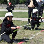 Na obrazku widać grupę dzieci ubranych w strażackie mundury, które ćwiczą używanie węży strażackich na trawiastym polu. Nad ich działaniami czuwają klasyczne ubrania w ubraniach.