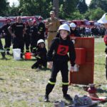 Na obrazku widać dzieci ubrane w strażackie mundury, które biorą udział w występach związanych z gaszeniem pożarów, występujących na trawiastym terenie. W tle możliwe jest wystąpienie kibiców oraz wozy strażackie.