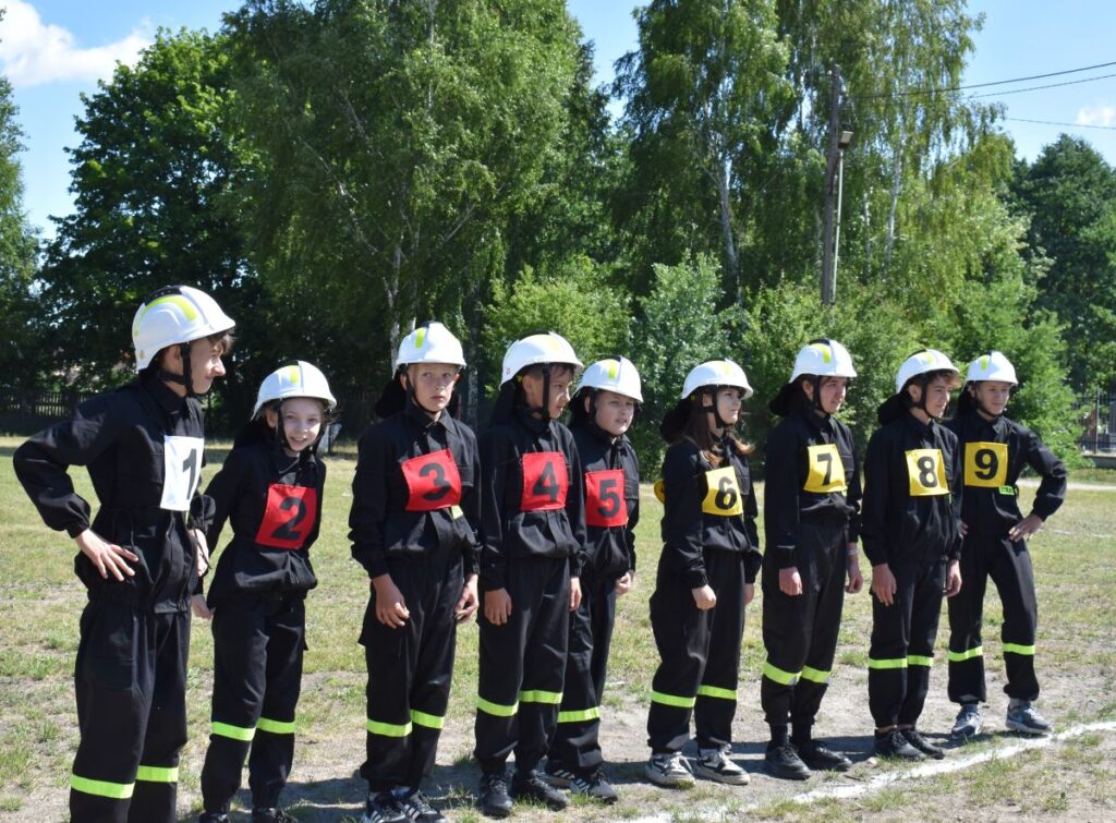 Na obrazku widać grupę młodych osób, ubranych w strażackie mundury i hełmy, każdy z nich ma na sobie numerowaną kamizelkę od 1 do 9. Stoją na trawie w szeregu, a za nimi rozpościera się tło z drzewami.