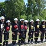 Na obrazku widać grupę młodych osób, ubranych w strażackie mundury i hełmy, każdy z nich ma na sobie numerowaną kamizelkę od 1 do 9. Stoją na trawie w szeregu, a za nimi rozpościera się tło z drzewami.