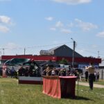 Na obrazku plenerowym z służb ratunkowych w mundurach, namiotach oraz torem dostępnym na trawiastym terenie. W tle znajduje się czerwony budynek z napisem „MAJA”, wokół którego rozmieszczony jest różnorodny sprzęt.