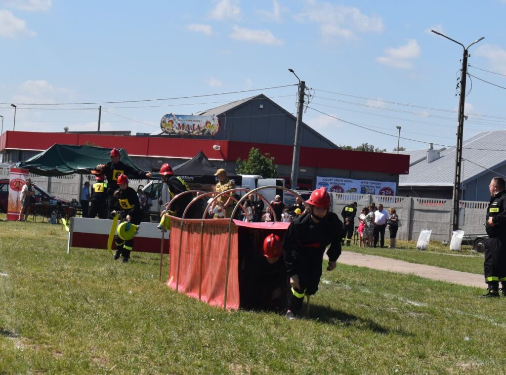 Strażacy, którzy są dostępni do udziału w ćwiczeniach na trawiastym polu, gdzie znajduje się czerwony budynek i zbiornik wodny. W tle wydarzenia obserwowani widzowie oraz inni członkowie mojej rodziny.