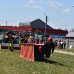 Strażacy, którzy są dostępni do udziału w ćwiczeniach na trawiastym polu, gdzie znajduje się czerwony budynek i zbiornik wodny. W tle wydarzenia obserwowani widzowie oraz inni członkowie mojej rodziny.