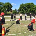 Na obrazku widać grupę osób, w tym dzieci, ubranych w strażackie mundury uczestniczących w ćwiczeniach przeciwpożarowych na trawiastej przestrzeni. Walczący ich standard oraz widzowie, którzy robią zdjęcia i uczestniczą w doświadczeniach ćwiczeń.