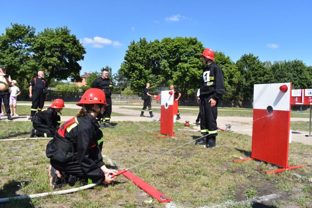 Na obrazku strażacy w urządzeniach treningowych uczestniczyli w ćwiczeniach, obsługując węże i strumienie wodne na czerwono-białe, umieszczone na trawiastym polu. W tle widoczni są drzewa i grupa obserwatorów.