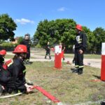 Na obrazku strażacy w urządzeniach treningowych uczestniczyli w ćwiczeniach, obsługując węże i strumienie wodne na czerwono-białe, umieszczone na trawiastym polu. W tle widoczni są drzewa i grupa obserwatorów.