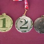 Na obrazku znajdują się trzy medale oznaczone numerami 1, 2 i 3, wyeksponowane na czerwonym tle. Każdy z medali jest podłączony do biało-czerwonej wstążki.