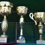 Na obrazku znajdują się trzy złote trofea z wygrawerowanymi tabliczkami, rozstrzygnięcie na ukrytym zielonym obrusem. W tle widoczny jest zielony namiot.