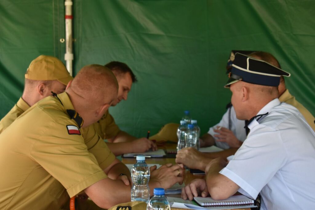 Dane dotyczące mężczyzn w mundurach wojskowych siedzących przy stole, na których znajdują się notatniki i butelki z wodą. W tle występuje zielone tło, a grupa jest zaangażowana w zasięgu.