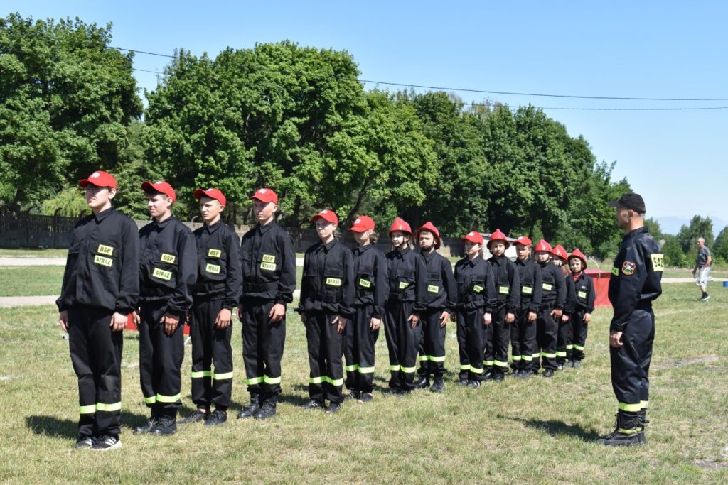 Na obrazku widać grupę umundurowanych osób w czapkach, stojących w szeregu na trawiastej powierzchni. W tle znajduje się drzewo, a przed osobą o dostępie do informacji.