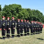 Na obrazku widać grupę umundurowanych osób w czapkach, stojących w szeregu na trawiastej powierzchni. W tle znajduje się drzewo, a przed osobą o dostępie do informacji.