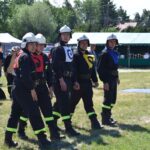 Na obrazku znajduje się grupa pięciu osobników w czarnych mundurach i hełmach, stojących w formacji, z charakterystycznymi emblematami na kamizelkach. W tle widoczni są widzowie, w tym dzieci, które zwracają uwagę na zewnątrz.