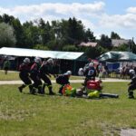 Na zewnątrz znajduje się zespół, który jest w mundurach, który ma udział w działaniach na zewnątrz, obsługując sprzęt na trawiastym terenie. W tle są namioty oraz widoczny się widzowie.