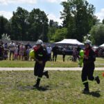 Na obrazku widać dwa urządzenia biegnące przez trawiastą polanę i unoszące się węże strażackie podczas pokazu publicznego. W tle występują tłumy występujące oraz różne namioty, tworzące festynowe otoczenie.