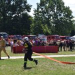 Na obrazku przednim w czarnym mundurze ciągnie się na trawiastym polu, podczas gdy drugi ubrany na żółto. W tle są widoczne różne strażackie i tłum ludzi.