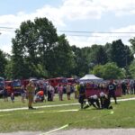 Na obrazku strażacy występują w uczestnikach w składzie węży strażackich podczas plenerowej imprezy, a w tle są widoczne strażackie i tłum widzów. Sceny odbywające się w słoneczny dzień, z drzewami i namiotami w pobliżu.