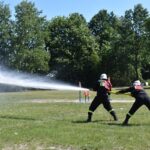 Na obrazku dwóch żołnierzy w czarnych żołnierzach i hełmach, którzy wysyłają strumień wody z wyczerpania podczas ćwiczeń. W tle znajduje się drzewo oraz trawiasta polana.