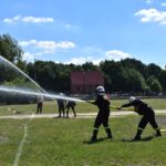 Na obrazku strażacy polewają wodę z węży podczas ćwiczeń na trawiastym polu, a w tle obserwującą ich ludzi. W widocznym widocznym budynku oraz drzewa.