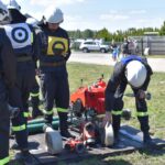 Na obrazku straży w mundurach wstępnych i węże wybuchowych oraz sprzętu na ziemi na zewnątrz. W tle są elementy krajobrazu, które powodują udostępnienie przestrzeni, może być miejscem ćwiczeń.