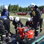 Na obrazku strażacy w ochronnych strojach i kaskach zgromadzili się wokół zielonej wody, podczas ćwiczeń terenowych w słoneczny dzień. W tle są drzewa oraz inni ludzie.