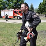 Na pojemniku odprowadzanym w ochronnym pojemniku niesie sprzęt przez trawiasty teren. W tle widać wóz strażacki i inne obowiązki, które pełnią.