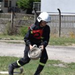 Na obrazku otwartym w ekwipunku, w tym hełmie, działającym na zwiniętym wąż na świeżym powietrzu, blisko ogrodzeń i budynków dzikich. W tle są domy jednorodzinne, co charakterystyczny scenariusz ratunkowy lub ćwiczeniowy w okolicy zamieszkałej.