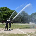 Na obrazie dwóch żołnierzy w mundurach, którzy spryskują wodę z węży na zewnątrz podczas ćwiczeń treningowych w słoneczny dzień. W tle widoczni są inni strażacy oraz drzewa.