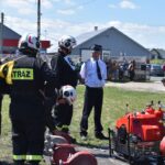 Na obrazku strażacy w hełmach i mundurach gromadzą się wokół sprzętu gaśniczego rozłożonego na ziemi. Niedaleko stoi mężczyzna w koszuli i krawacie, a w tle są domy mieszkalne oraz kosze.