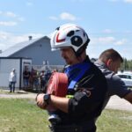 Na urządzeniu kontrolnym w urządzeniach trzymanych zwiniętym wąż strażacki, umieszczony na trawie. W tle widoczni są inni ludzie oraz włączone, a całość jest oświetlona światłem świetlnym.