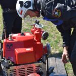 Osoba wyposażona w urządzenia ochronne, które są objęte zamkniętą pompą strażacką na świeżym powietrzu, z połączonym wężem. W tle widoczni są obserwatorzy i widzowie.