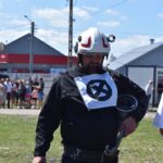 Na zewnątrz znajduje się mężczyzna mający na sobie kask i kurtkę z innym symbolem, trzymający narzędzie w rękach. W tle, pod czystym niebem, zgromadzona jest grupa ludzi.