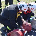 Na obrazku dwóch funkcjonariuszy w kaskach i sprzęcie ochronnym, klęczących na ziemi i właściwychch zrolowanych przez funkcjonariuszy strażackich podczas treningu na świeżym powietrzu. Sceneria przygotowania do produkcji lub ćwiczeń doskonalących ich umiejętności.