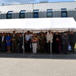 Na obrazku jest grupa osób, w skład której wchodzą jednostki mundurowi, jak i cywile, stojące pod białym zadaszeniem przed bezprzewodowym wyłącznikiem. Niektórzy uczestnicy trzymają kwiaty.