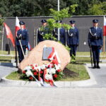 Na obrazku znajduje się grupa honorowych wartowników w mundurach, stojących na baczność wokół pamiątkowego kamienia umieszczonego nad głową i wstęgami. Po obu pomnikach są polskie flagi.