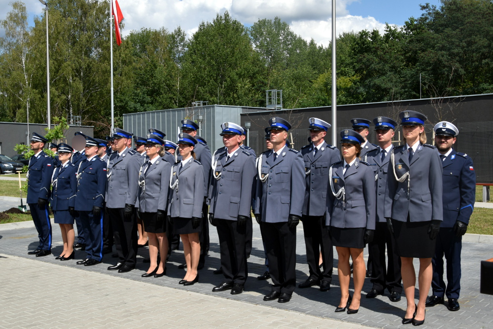 Na obrazku widać grupę umundurowanych oficerów stojących w zagrożeniu na świeżym powietrzu, a w tle znajduje się dach i samochód. Kilku z nich trzyma flagi, a pogoda wydaje się być słoneczna i bezchmurna.