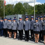 Na obrazku widać grupę umundurowanych funkcjonariuszy stojących w formacji na zewnątrz. W tle znajduje się drzewo oraz maszt z podniesionymi flagami.