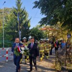 20240801_170302 Na obrazku widocznym grupy osób, w tym przeznaczonych w mundurach, przechodzących chodnikiem. Na czele pochodu znajdują się osoby unoszące się z ozdobną wstążką, a tłem są drzewa i budynek.