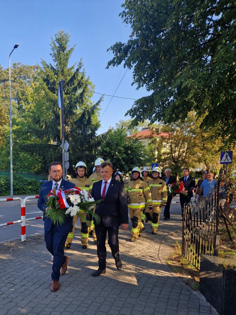 Na obrazku widocznym grupy osób, w tym przeznaczonych w mundurach, przechodzących chodnikiem. Na czele pochodu znajdują się osoby unoszące się z ozdobną wstążką, a tłem są drzewa i budynek.